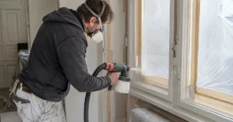 Heimwerker sprüht einen abgeklebten Holzfensterrahmen mit einem elektrischen Farbsprühsystem
