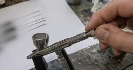 Hand stellt die Nadelbegrenzung einer Airbrushpistole ein, daneben feine Probelinien auf Papier